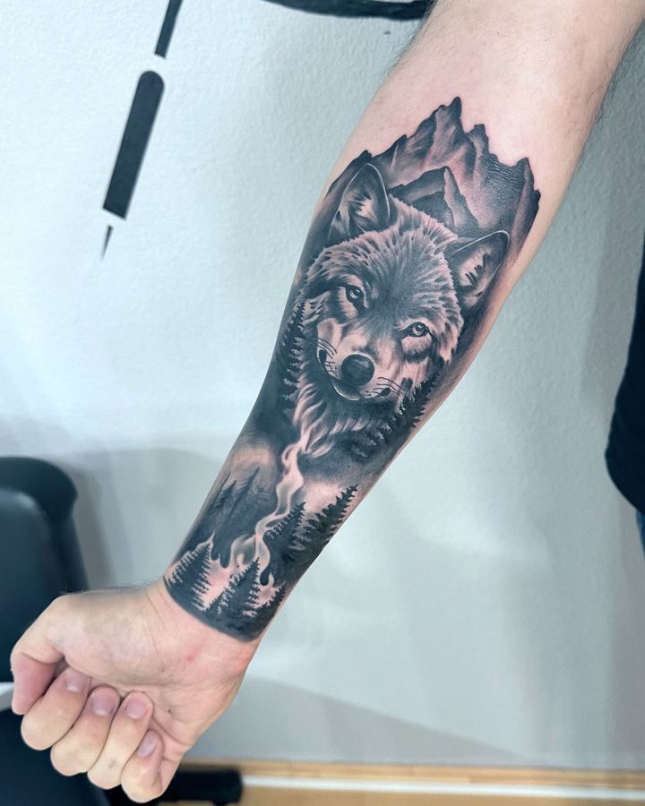Black-and-Grey-Tattoo eines Wolfs vor Bergen und Wald auf dem Unterarm